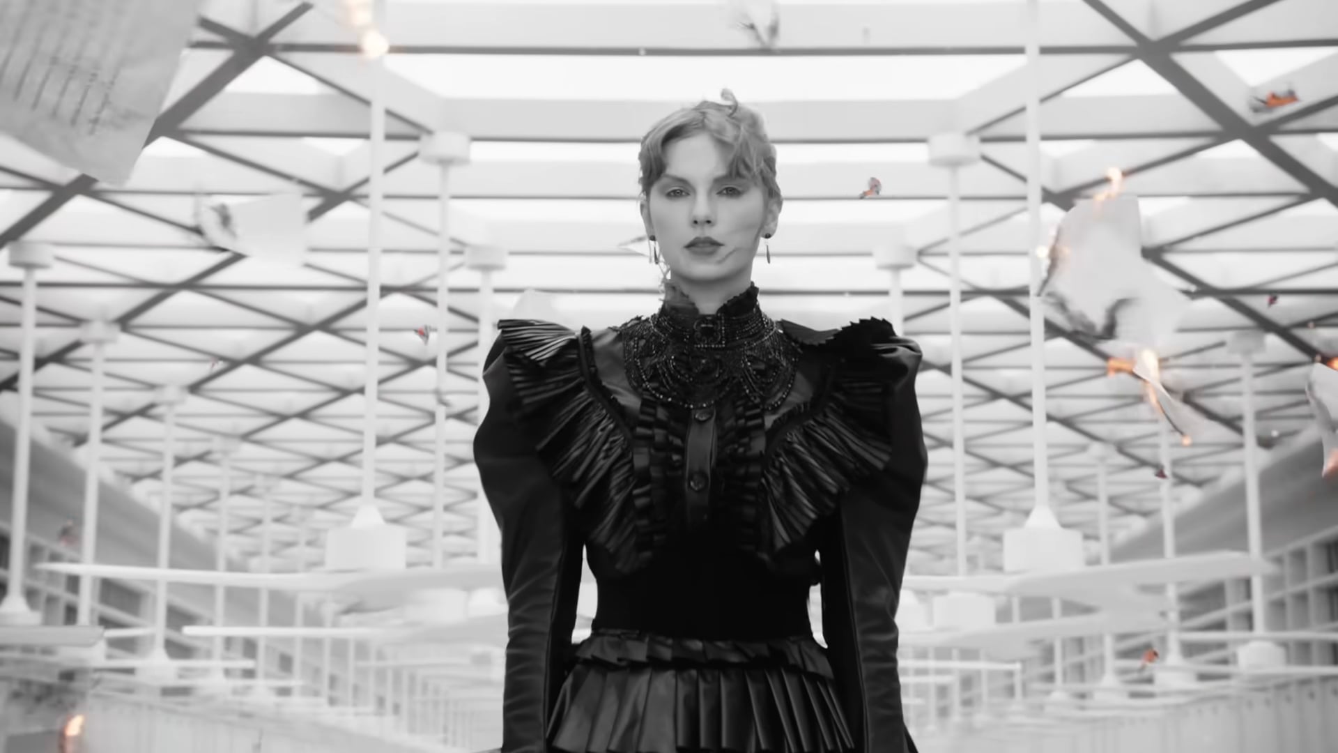 Taylor Swift in a black dress standing in the middle of a room with papers flying around it